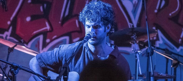 Un giovane ragazzo con una chitarra acustica che canta in modo intenso e concentrato su un palco, in un contesto urbano con graffiti su un muro sullo sfondo.